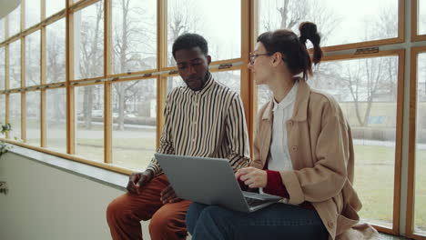 Diverse-Man-and-Woman-Using-Laptop-and-Speaking-on-Window-Sill